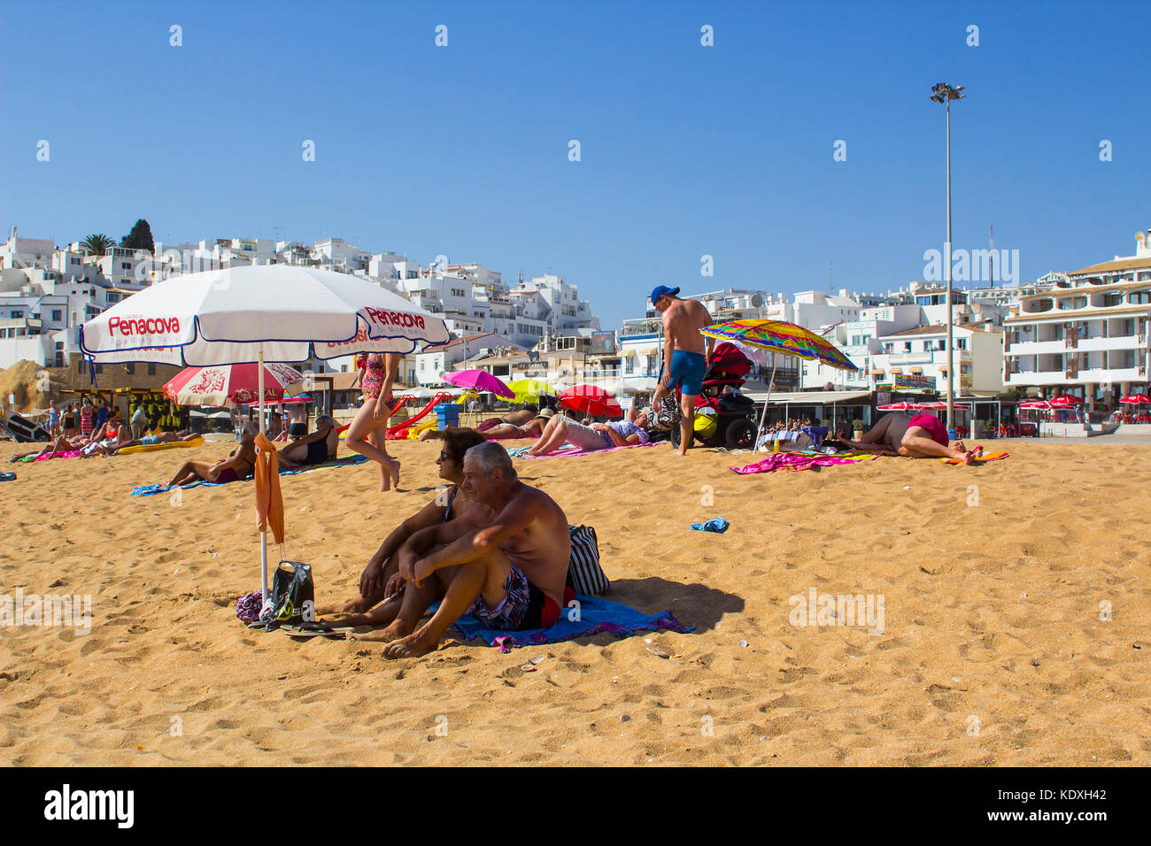 An elderly couple sitting in the shade of a sun brolly on the sandy beach in Albuferia in Portugal Stock Photo