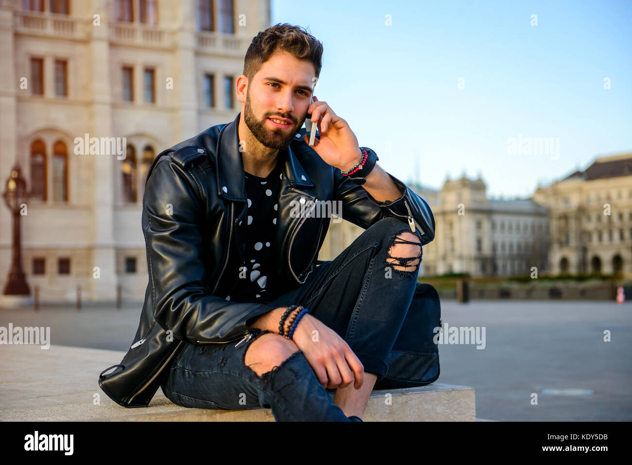 A handsome young hipster man sitting on the stairs on a square while ...