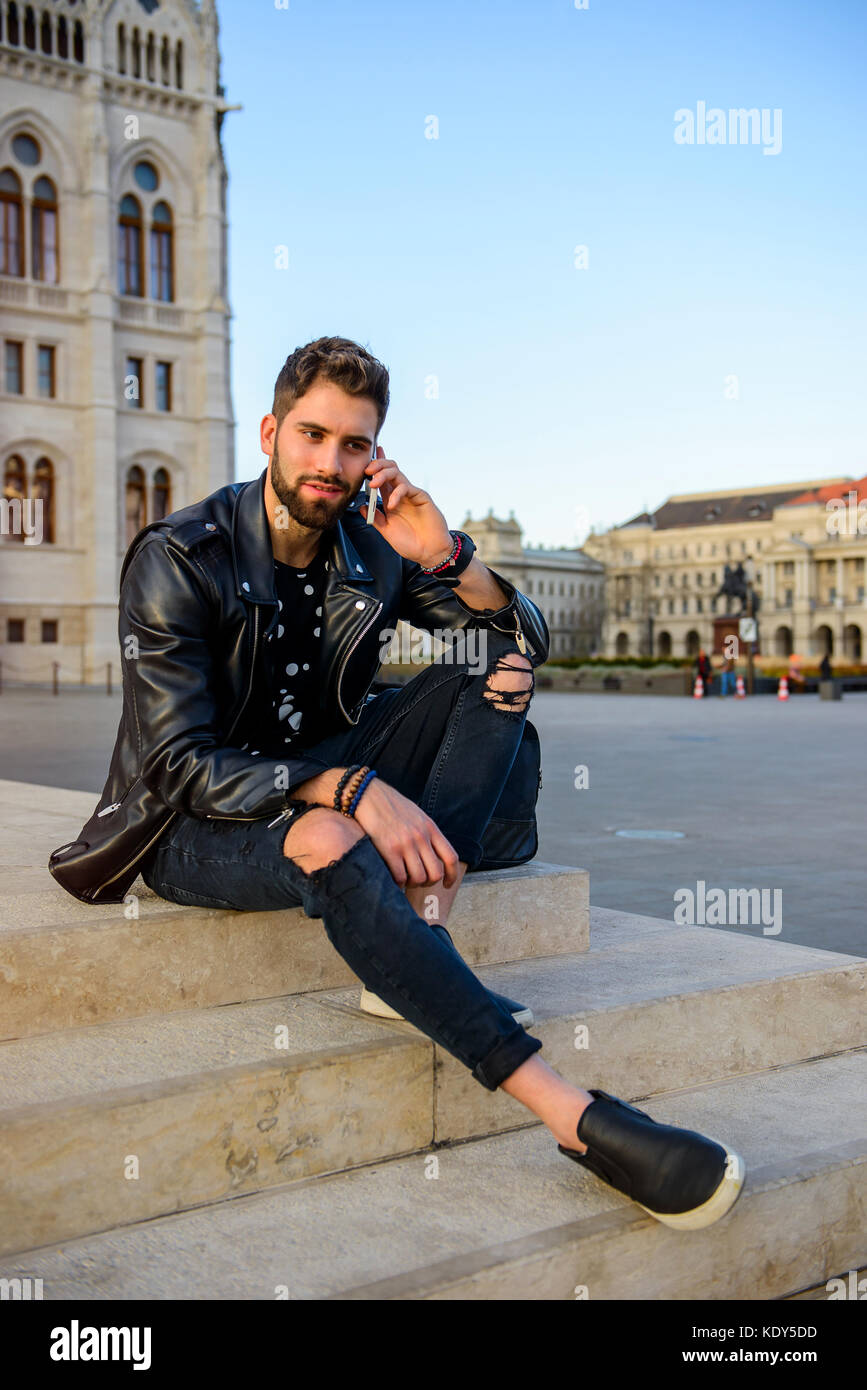 A handsome young hipster man sitting on the stairs on a square while ...