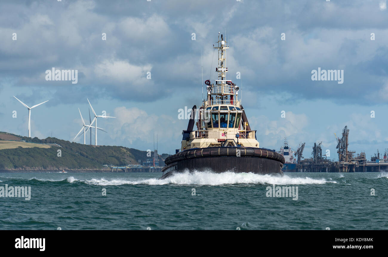 A powerful tug creates a huge bow wave as it heads past the Valero oil terminal with wind turbines in the background, slowing down for a passing yacht Stock Photo