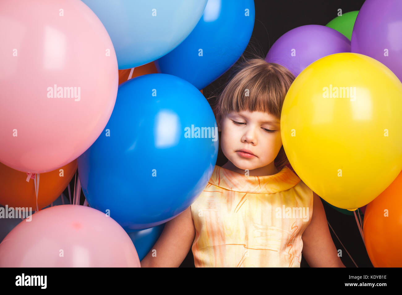 Portrait of sad Caucasian blond little girl with colorful balloons over black background Stock Photo