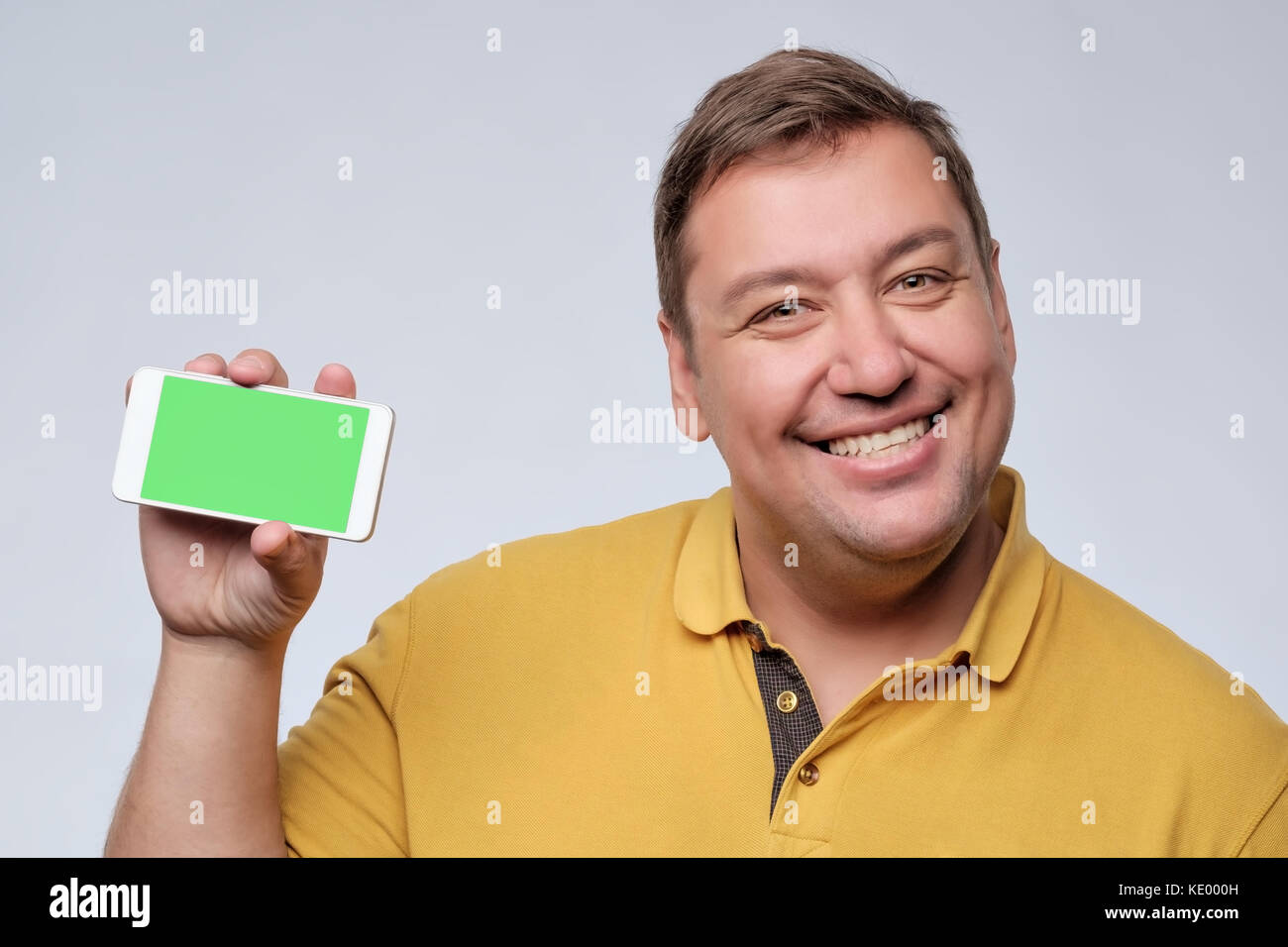 Fat man in a yellow shirt pleased with himself holding a phone with green screen. Stock Photo