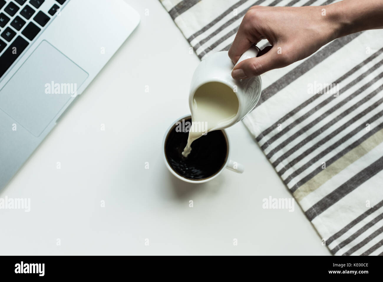 Person pouring milk from jug into cup of black coffee on white working