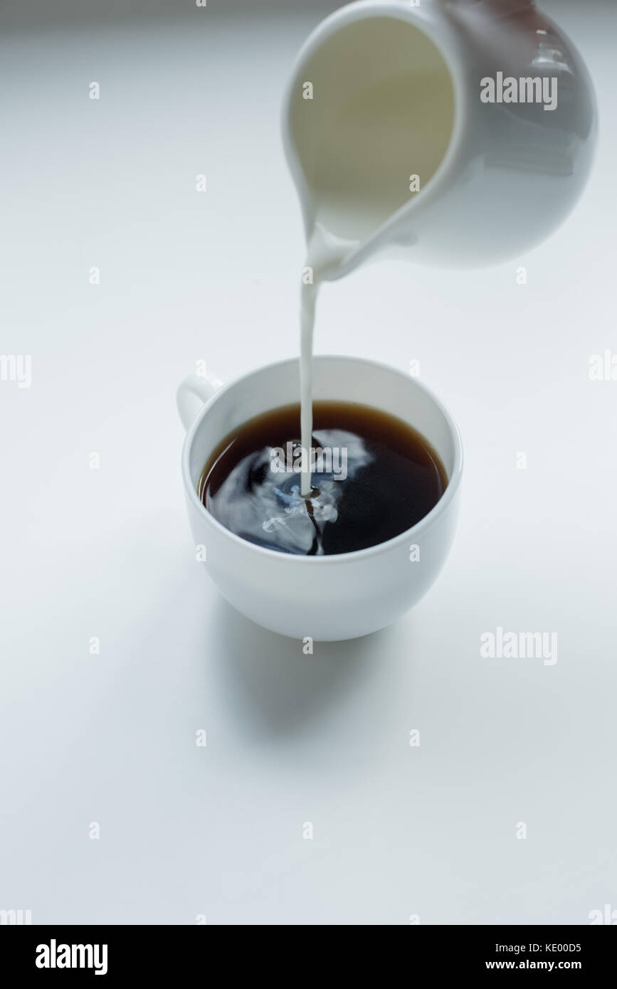 Person pouring milk from jug into cup of black coffee on white table