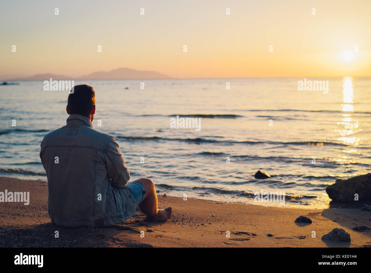 A young guy is sitting on the beach of the sea and looking at the ...