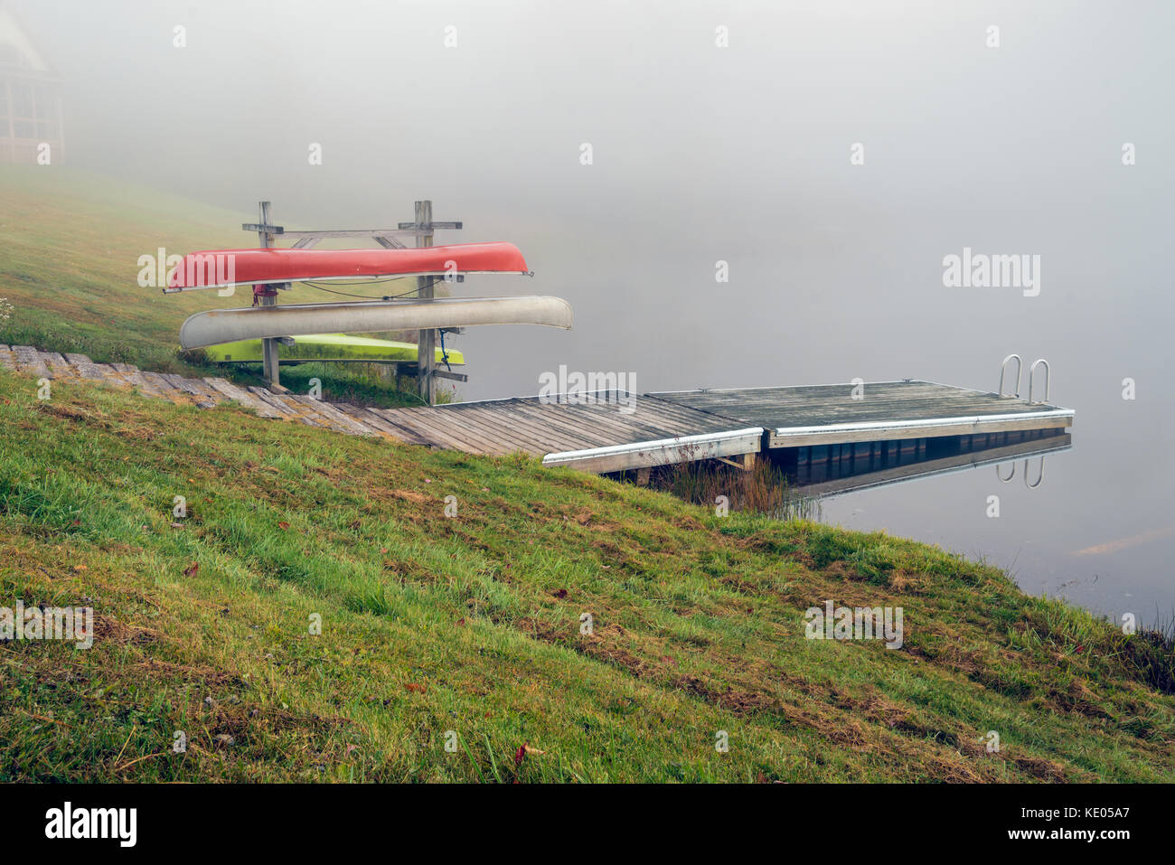 Three canoes on a rack by the side of a calm lake on a foggy morning in the fall. Stock Photo