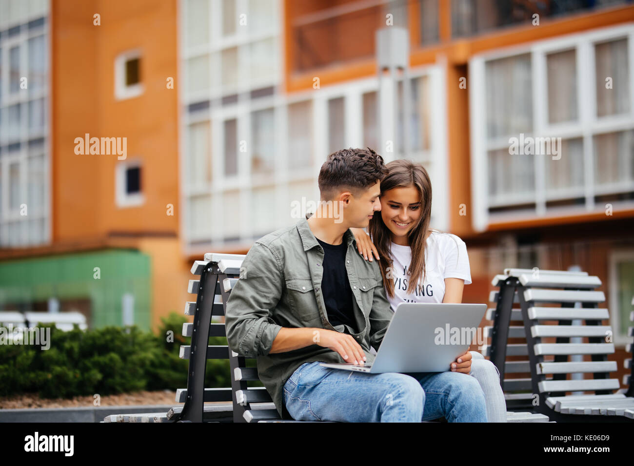 Happy couple using laptop while sitting on bench in park and smiling ...