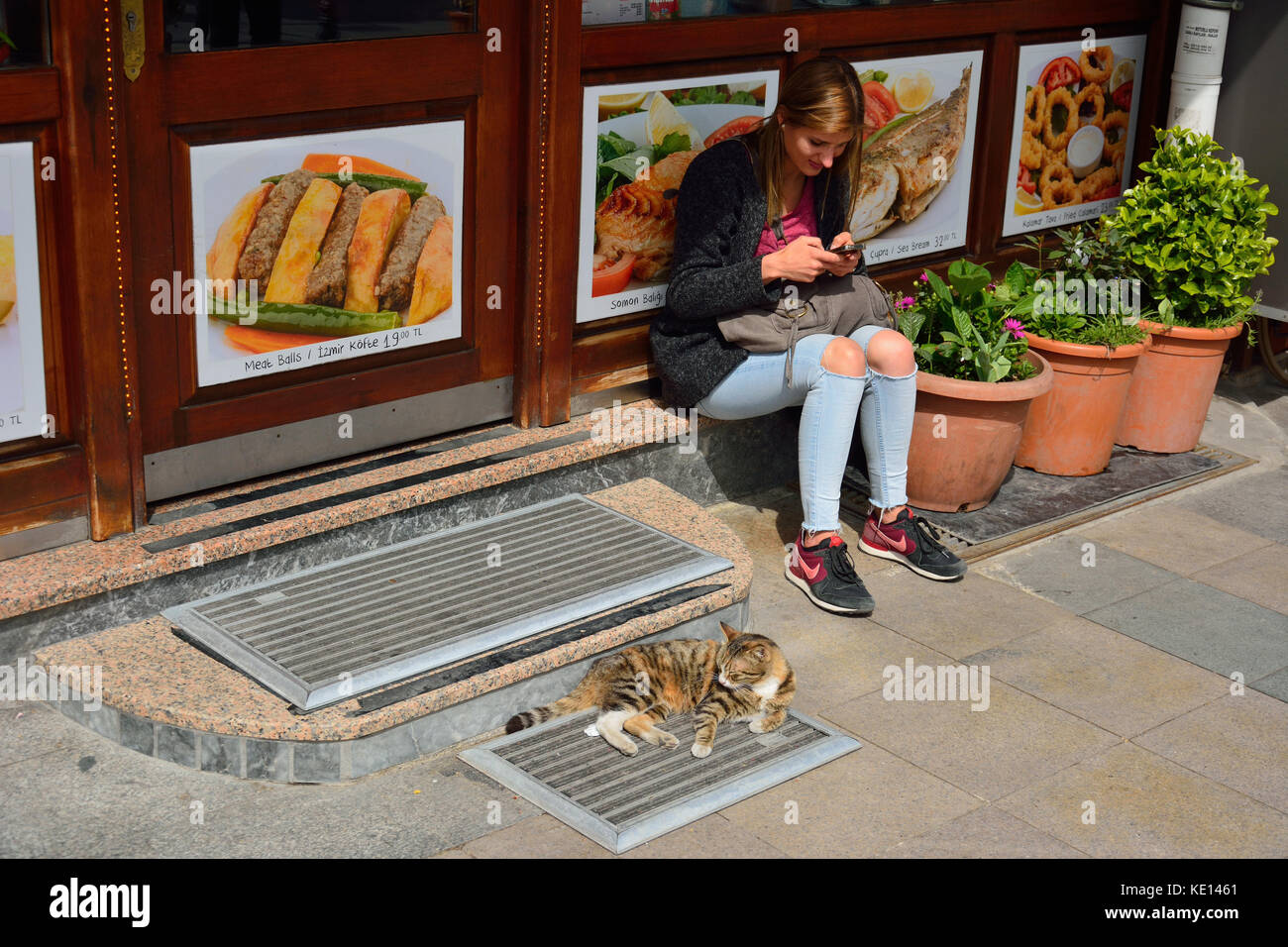 Istanbul, Turkey - April 22, 2017. Street scene in Istanbul, with girl sitting and holding a smartphone and cat lying in front of kebab shop in Istanb Stock Photo