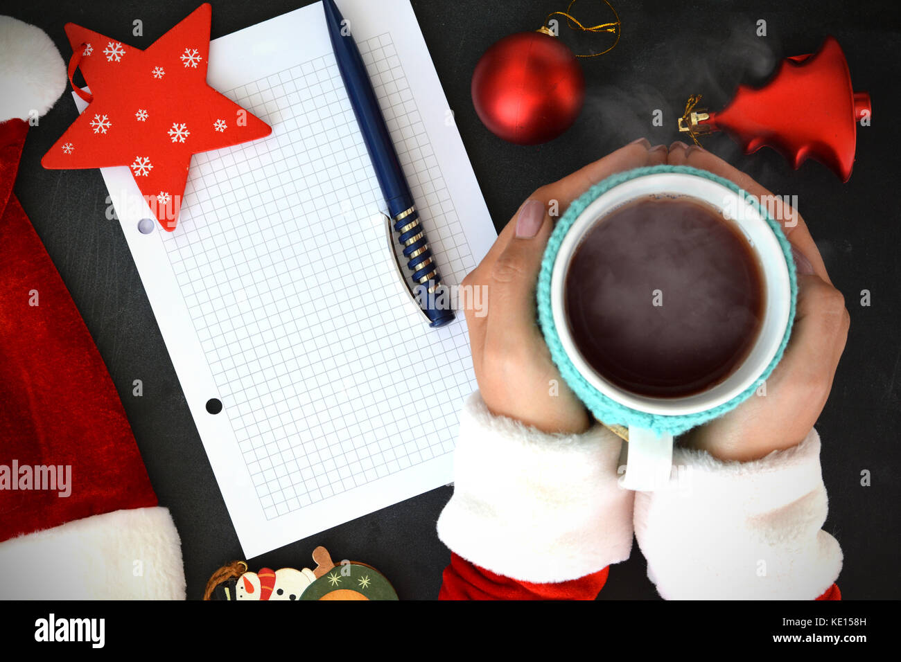 Woman in Santa’s clothes holding a cup of hot tea on Christmas background, near an empty wish list Stock Photo