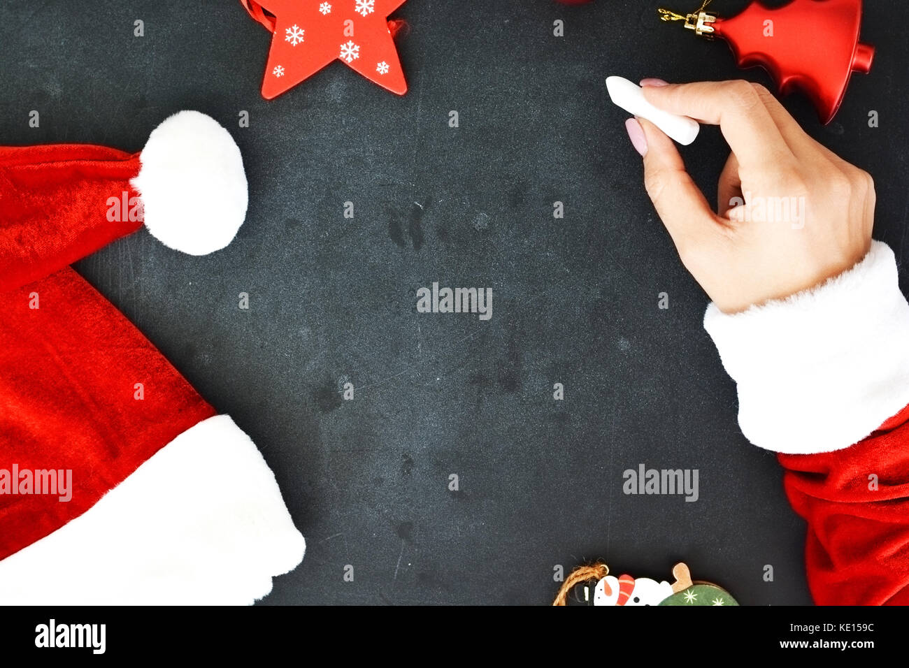 Christmas background with woman hands dressed in Santa’s costume writing on empty blackboard with seasonal decorations Stock Photo