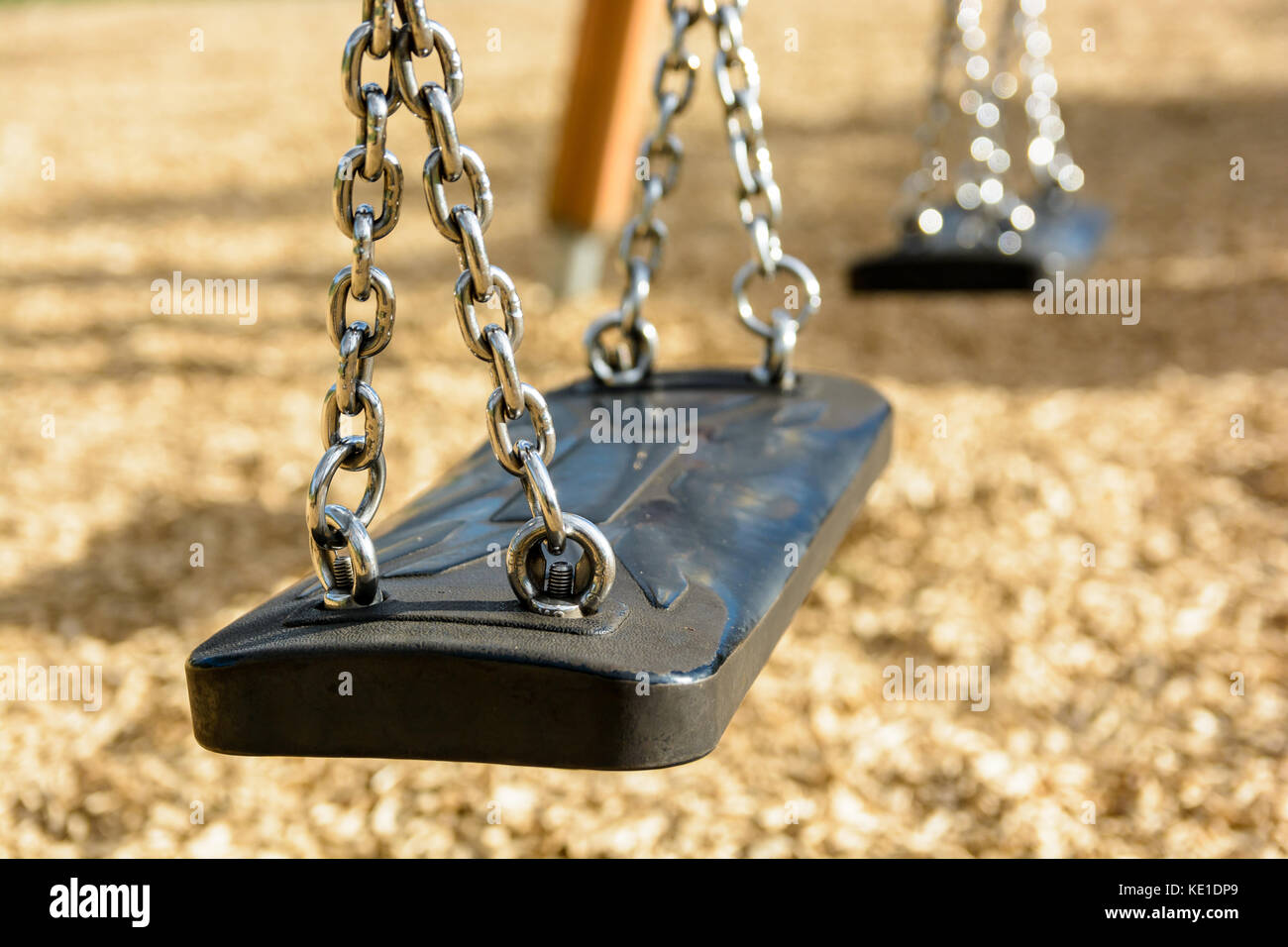 Close-up view of a still child's swing in black plastic in a wood chips covered playground with chrome chains and drop shadow. Stock Photo