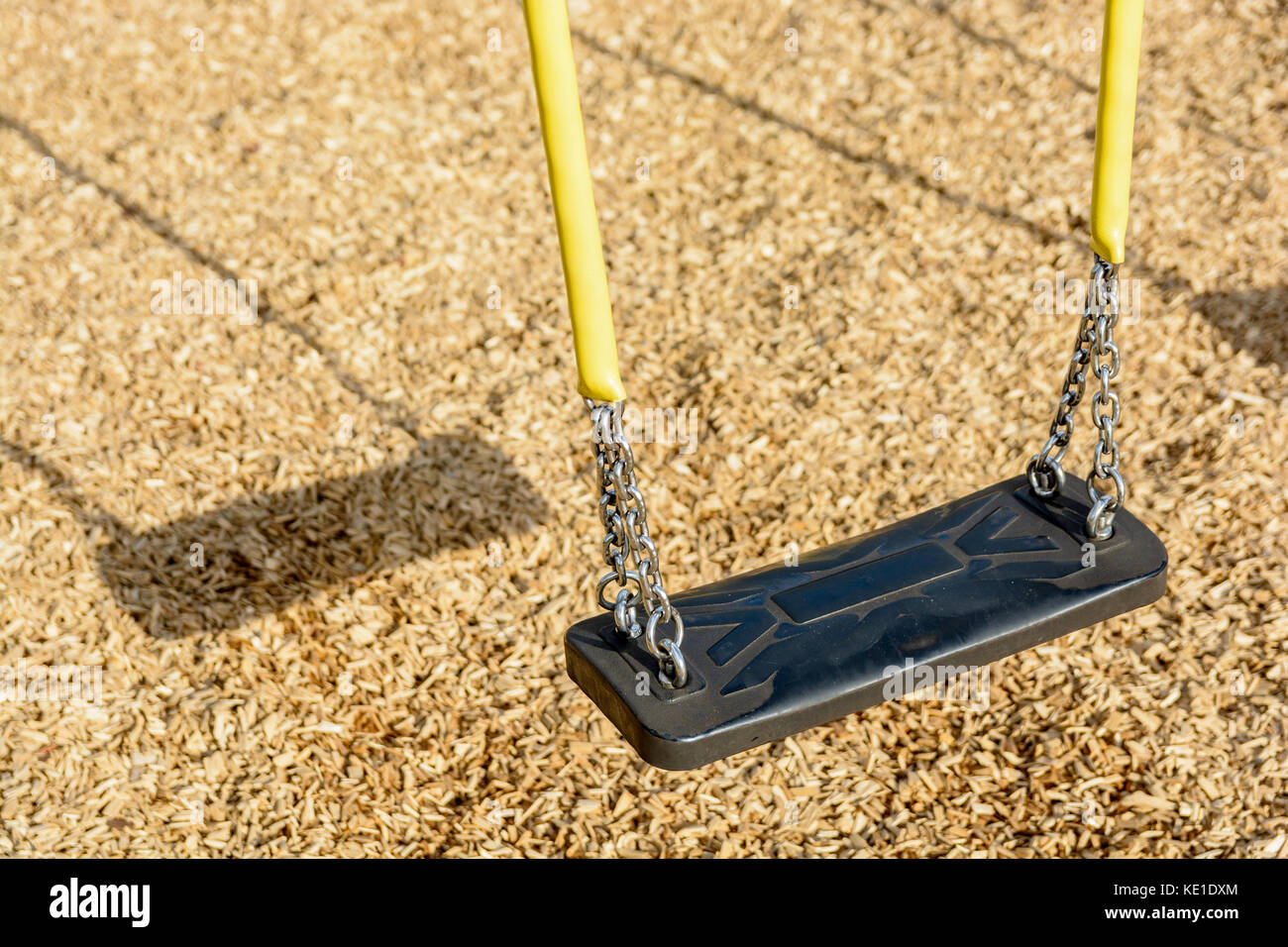 A still child's swing in black plastic in a wood chips covered playground with chrome chains in a yellow plastic sleeve and drop shadow. Stock Photo
