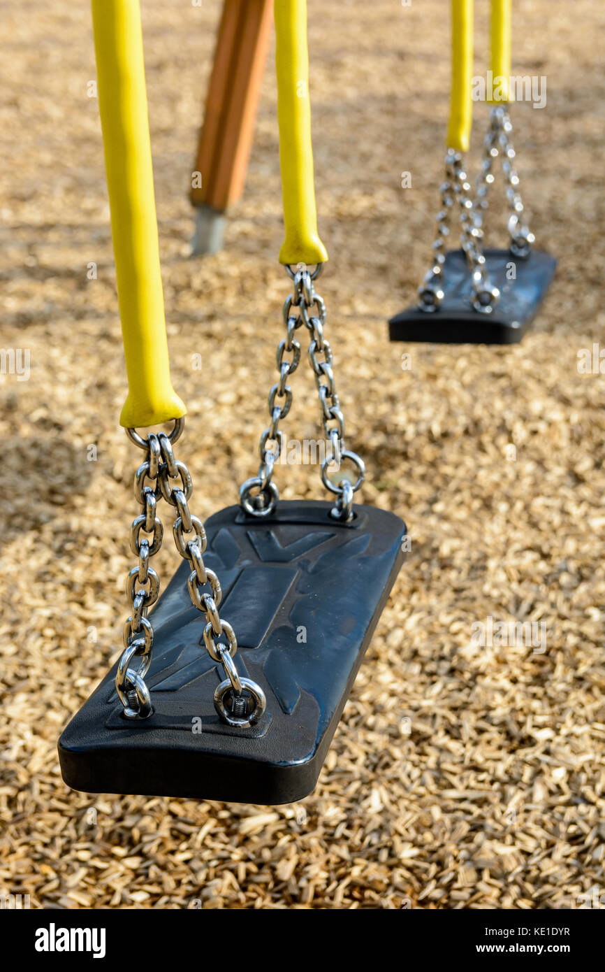 A still child's swing in black plastic in a wood chips covered playground with chrome chains in a yellow plastic sleeve. Stock Photo