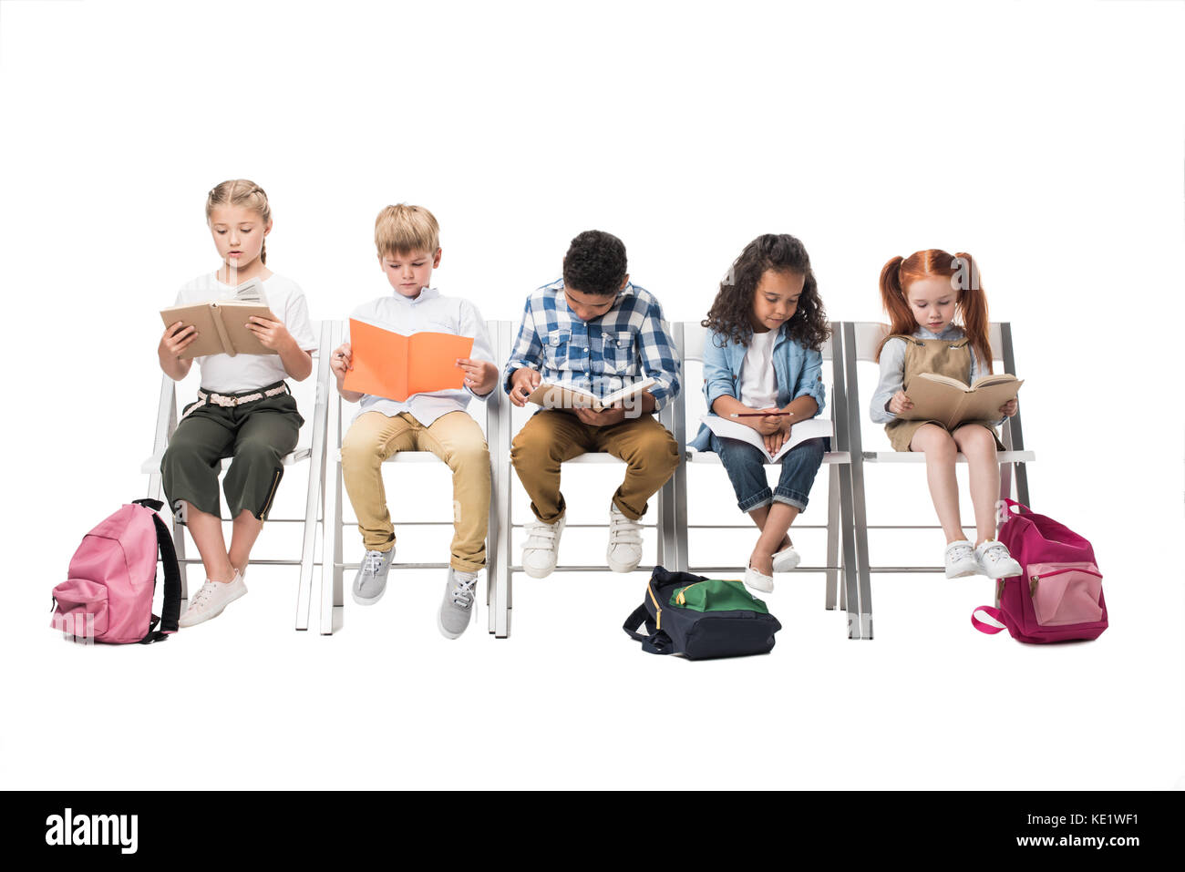 multiethnic kids reading books while sitting on chairs isolated on white Stock Photo