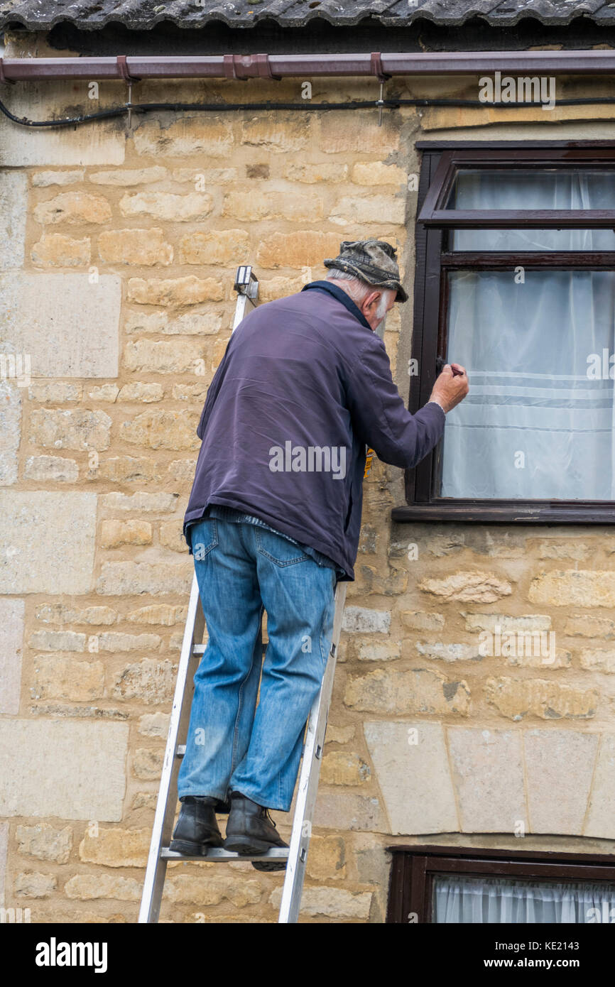 Sprightly old man, wearing hat, jacket and denim jeans, up a ladder, painting a window frame on the upper storey of a house. Langtoft, England, UK. Stock Photo