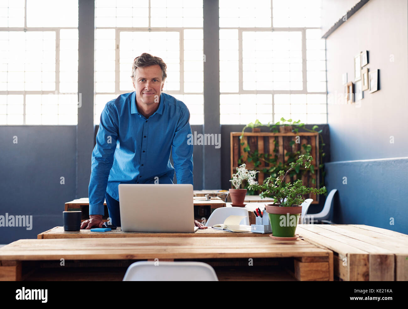 Portrait of a smiling businessman leaning confidently over a laptop on his desk in a large modern office working online with a laptop Stock Photo