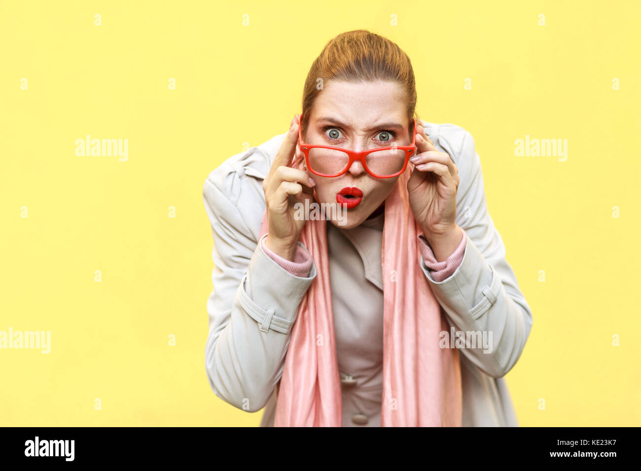 Redhead woman wearing coat, opening mouths widely, having surprised shocked looks.  Studio shot, isolated on yellow wall Stock Photo