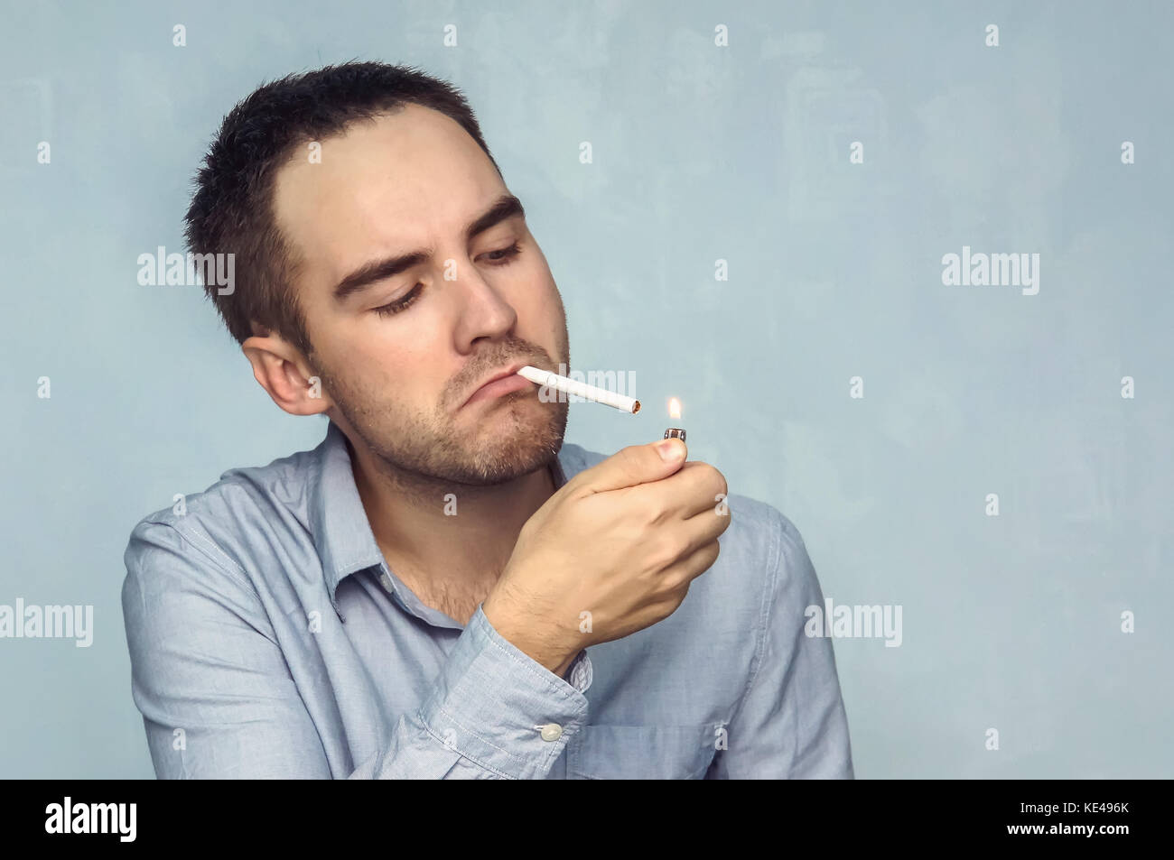 Young man lighting cigarette. Businessman in a shirt he lights a cigarette from the flame of the lighter on a blue background. Stock Photo