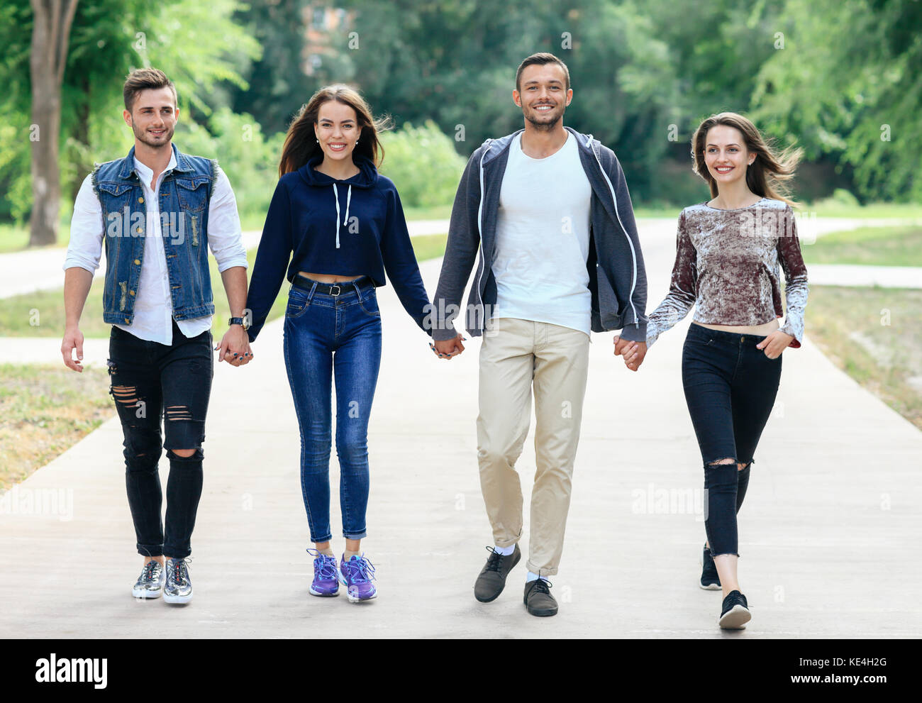 Full-length portrait of young friends holding hands walking in park. Four people happy looking at camera. Two guys and two girls in stylish casual clo Stock Photo