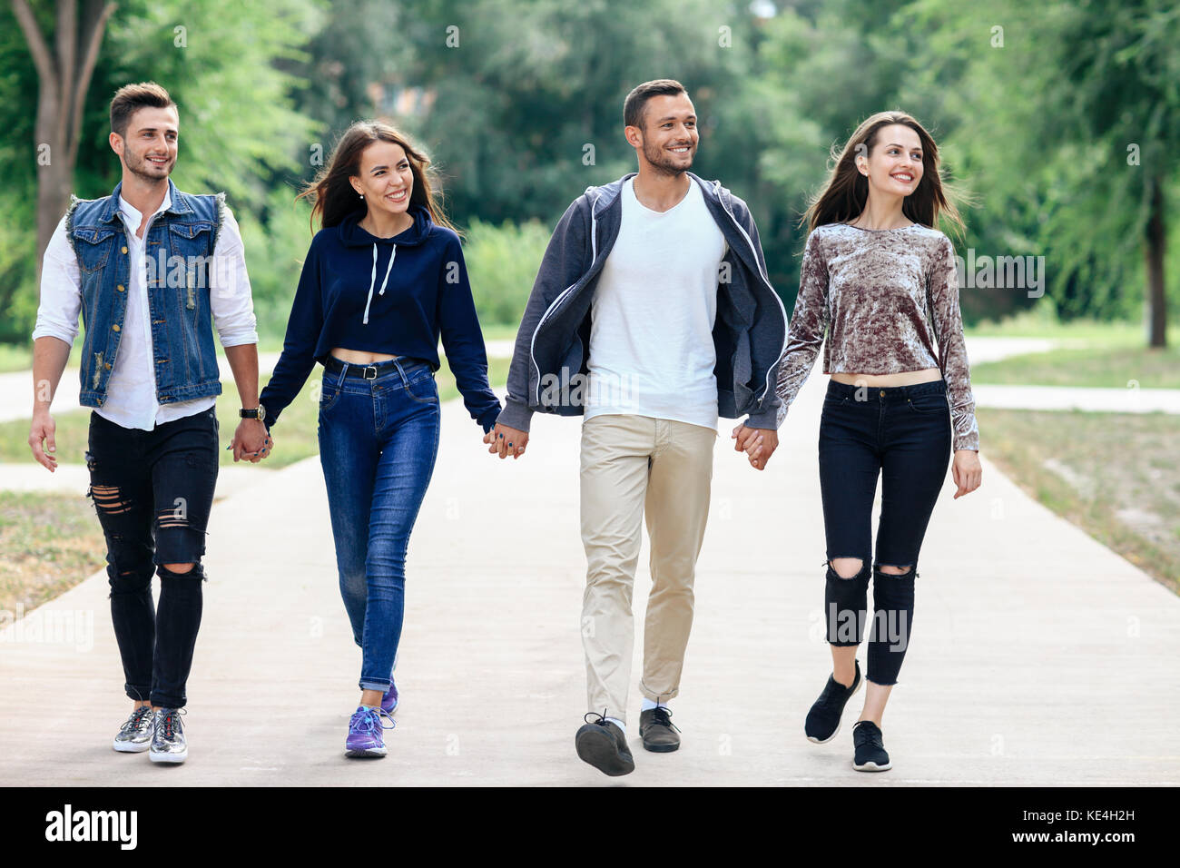 Full-length portrait of young friends holding hands walking in park. Four people happy looking at camera. Two guys and two girls in stylish casual clo Stock Photo