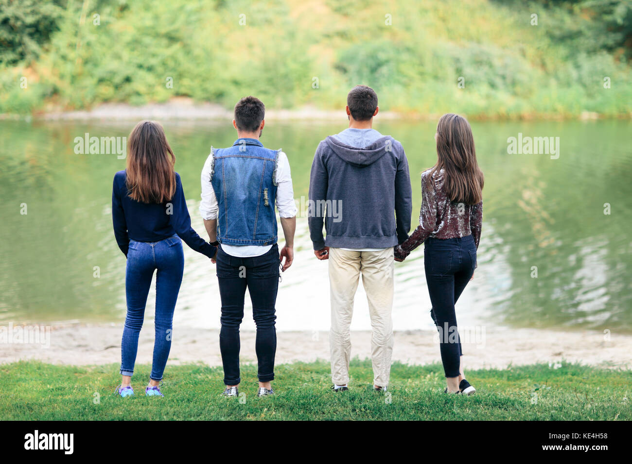 Two love couples standing by lake in park back view. Romantic rest of four young people in nature, two girls and two guys looking at calm water. Conte Stock Photo