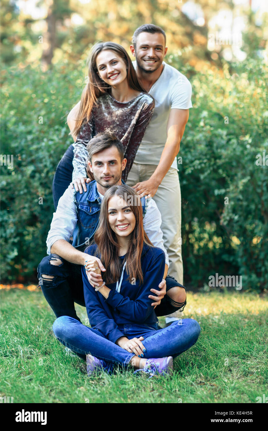 Men and women on nature. Four happy friends smiling and looking at camera. Young girls and guys sitting and standing on grass. Portrait of cheerful pe Stock Photo