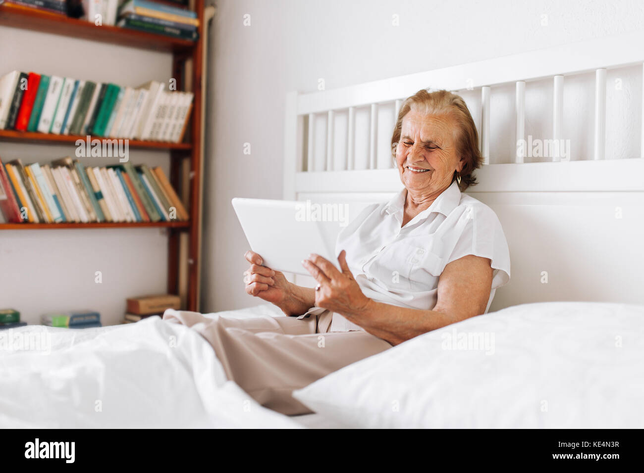 Elderly woman sitting comfortably on bed and using her digital tablet Stock Photo