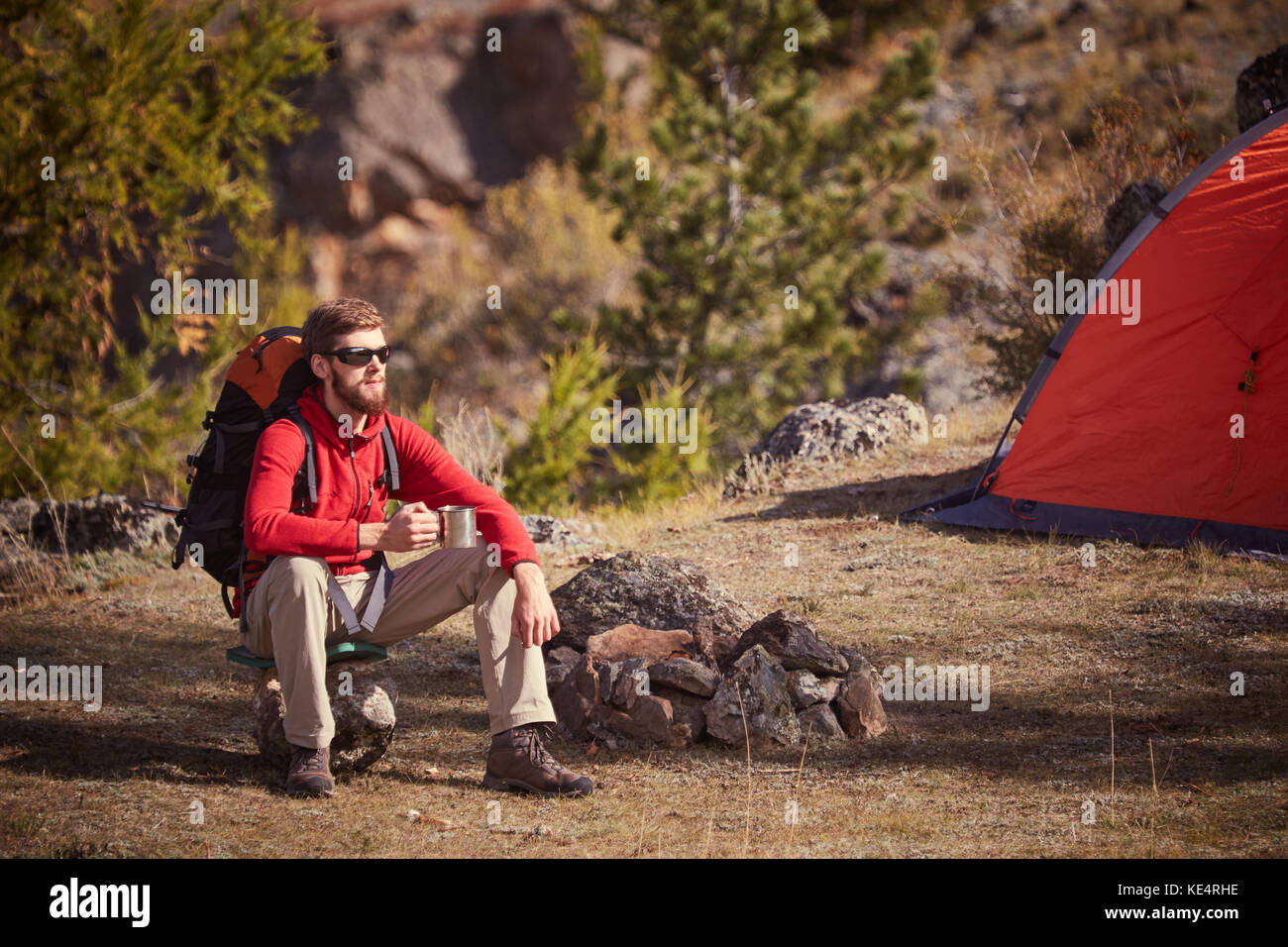 Backpacker having rest near orange tent with cup in hand. Stock Photo