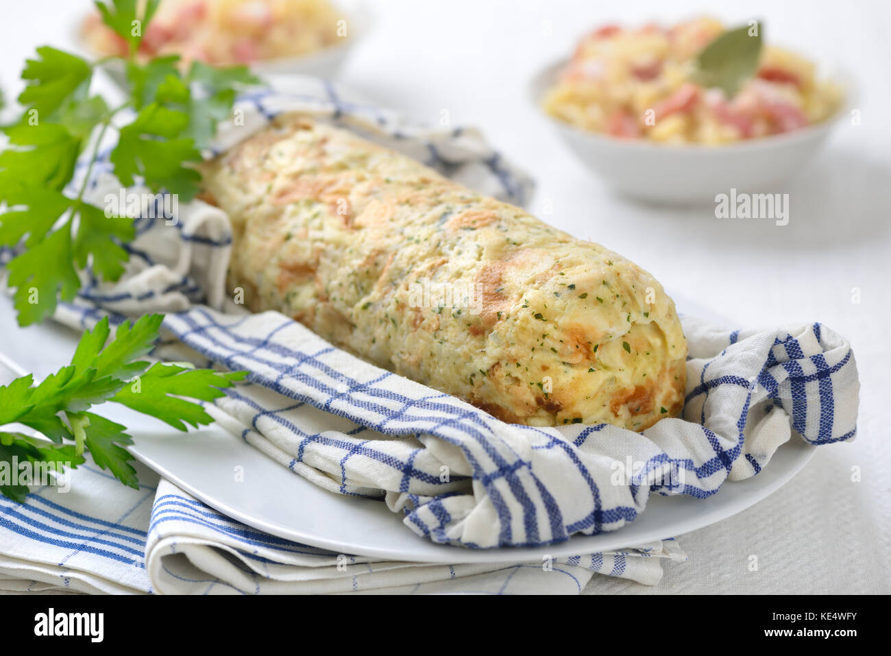 Uncut Bavarian bread dumpling cooked in a rolled up tea towel, sauerkraut with bacon in the background Stock Photo