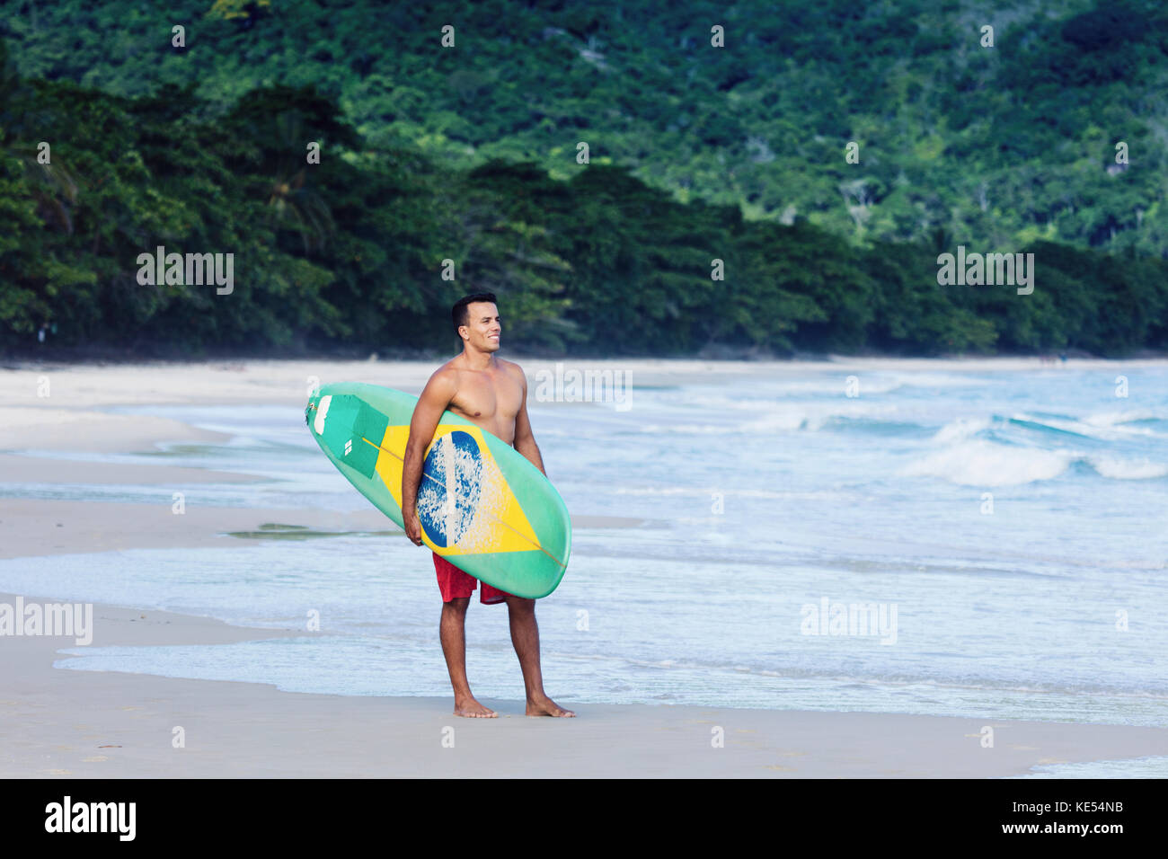 Surfer on pristine beach in Rio de Janeiro state, carrying his board, which shows a Brazilian flag Stock Photo