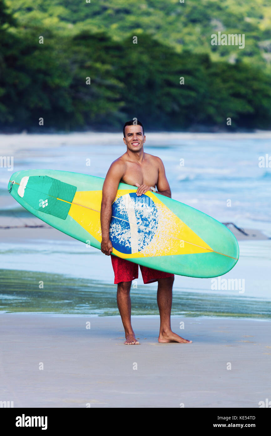 Surfer on pristine beach in Rio de Janeiro state, carrying his board, which shows a Brazilian flag Stock Photo