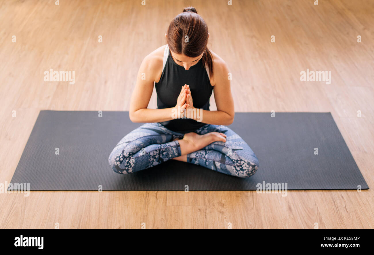 High angle view of young woman working out at home, doing yoga exercise on mat. Female sitting half Lotus pose. Ardha padmasana yoga pose. Stock Photo
