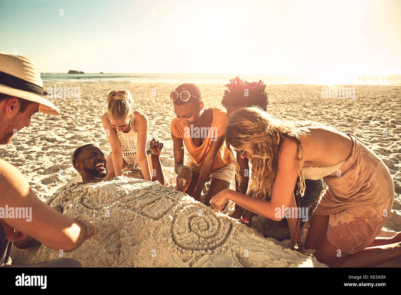 Young friends burying and drawing friend in sand on sunny summer beach Stock Photo