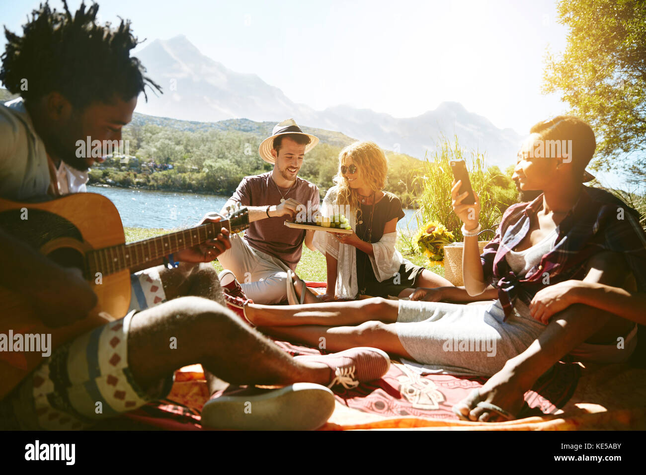Young friends hanging out, playing guitar and enjoying picnic at sunny summer riverside Stock Photo