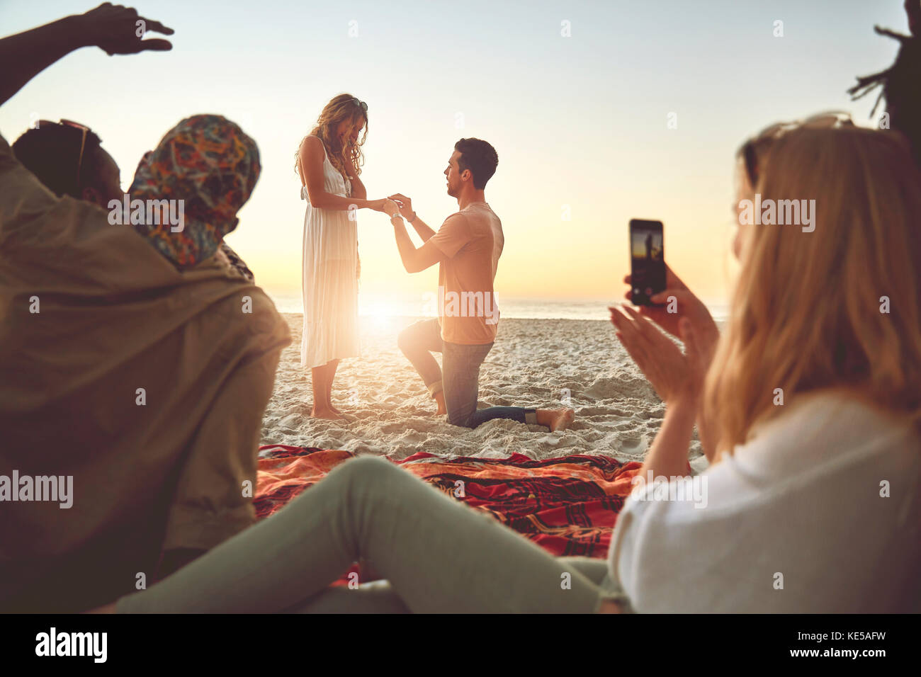 Young man proposing to woman on sunny summer beach with friends Stock Photo