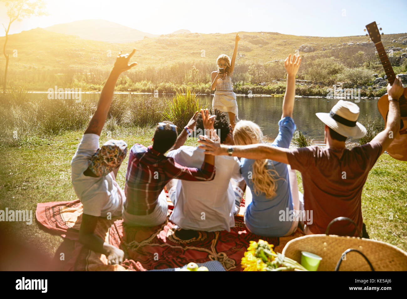 Woman with camera phone photographing friends with arms raised at sunny summer riverside Stock Photo