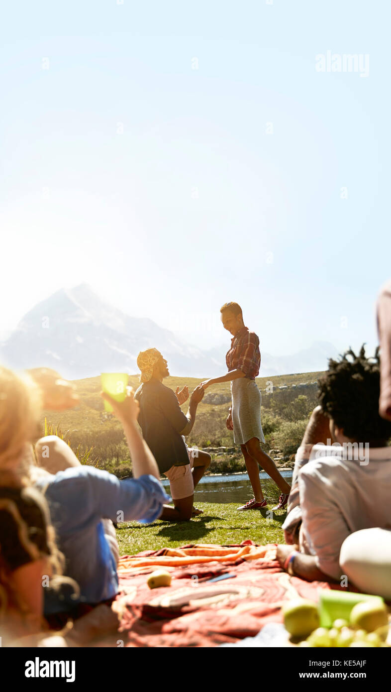Friends watching young man proposing to woman at sunny summer picnic Stock Photo