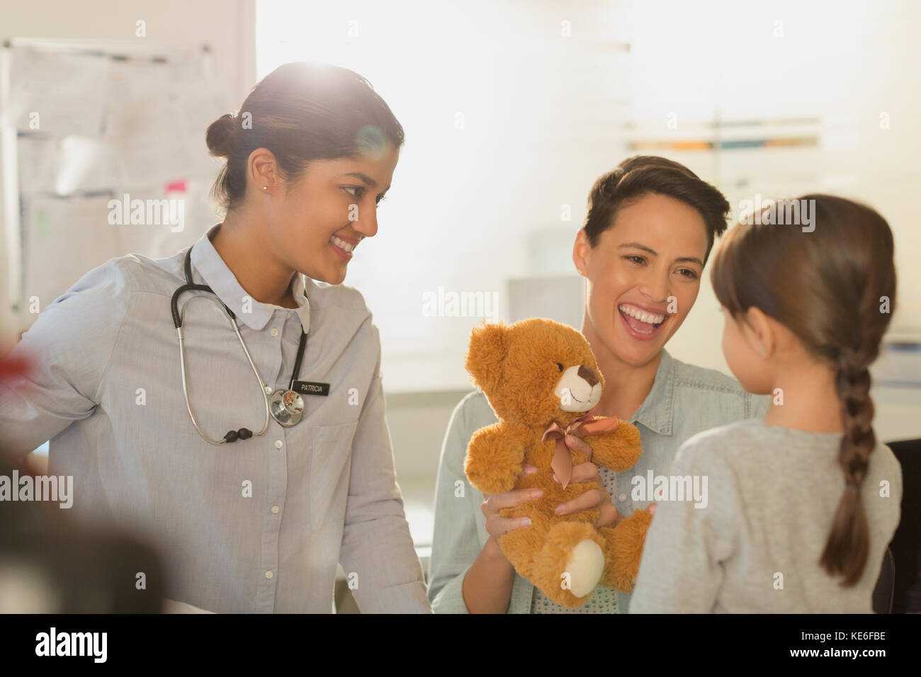 Smiling female pediatrician and mother showing teddy bear to girl patient in examination room Stock Photo