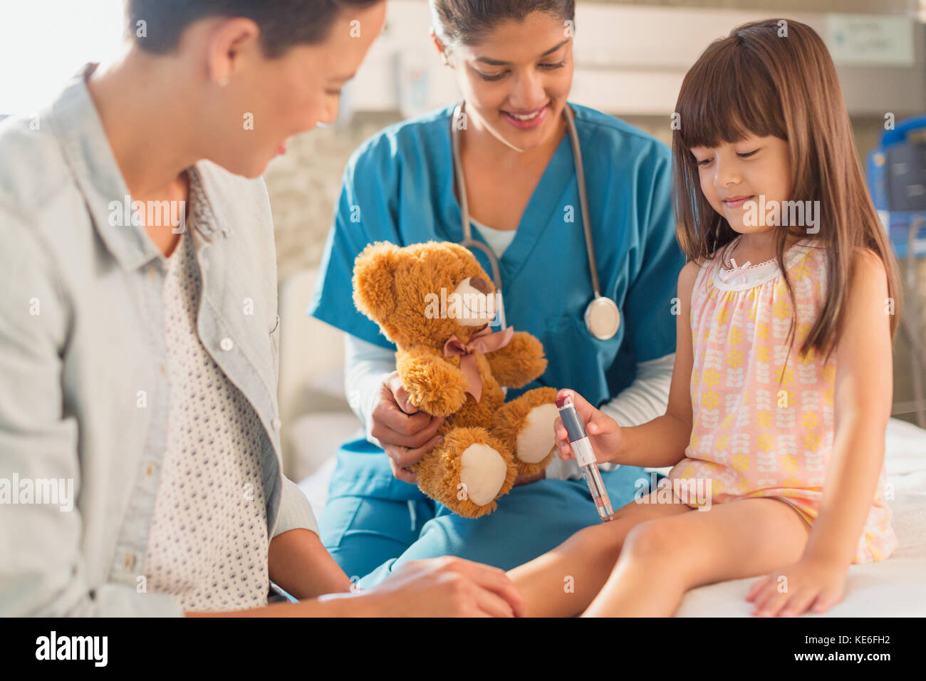 Female nurse with teddy bear watching girl patient using insulin pen in hospital room Stock Photo