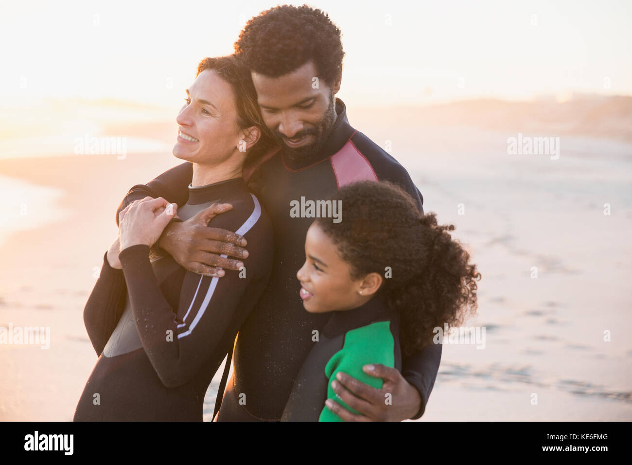 Affectionate multi-ethnic family in wet suits hugging on sunset beach Stock Photo