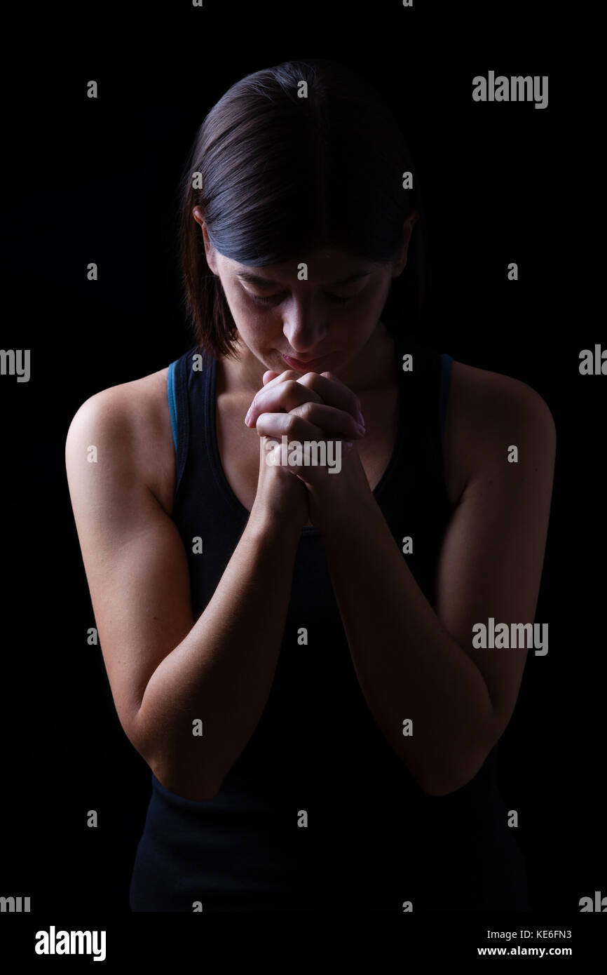 Faithful athletic woman praying, with hands folded in worship to god, head down and eyes closed in religious fervor, on low key black background. Stock Photo