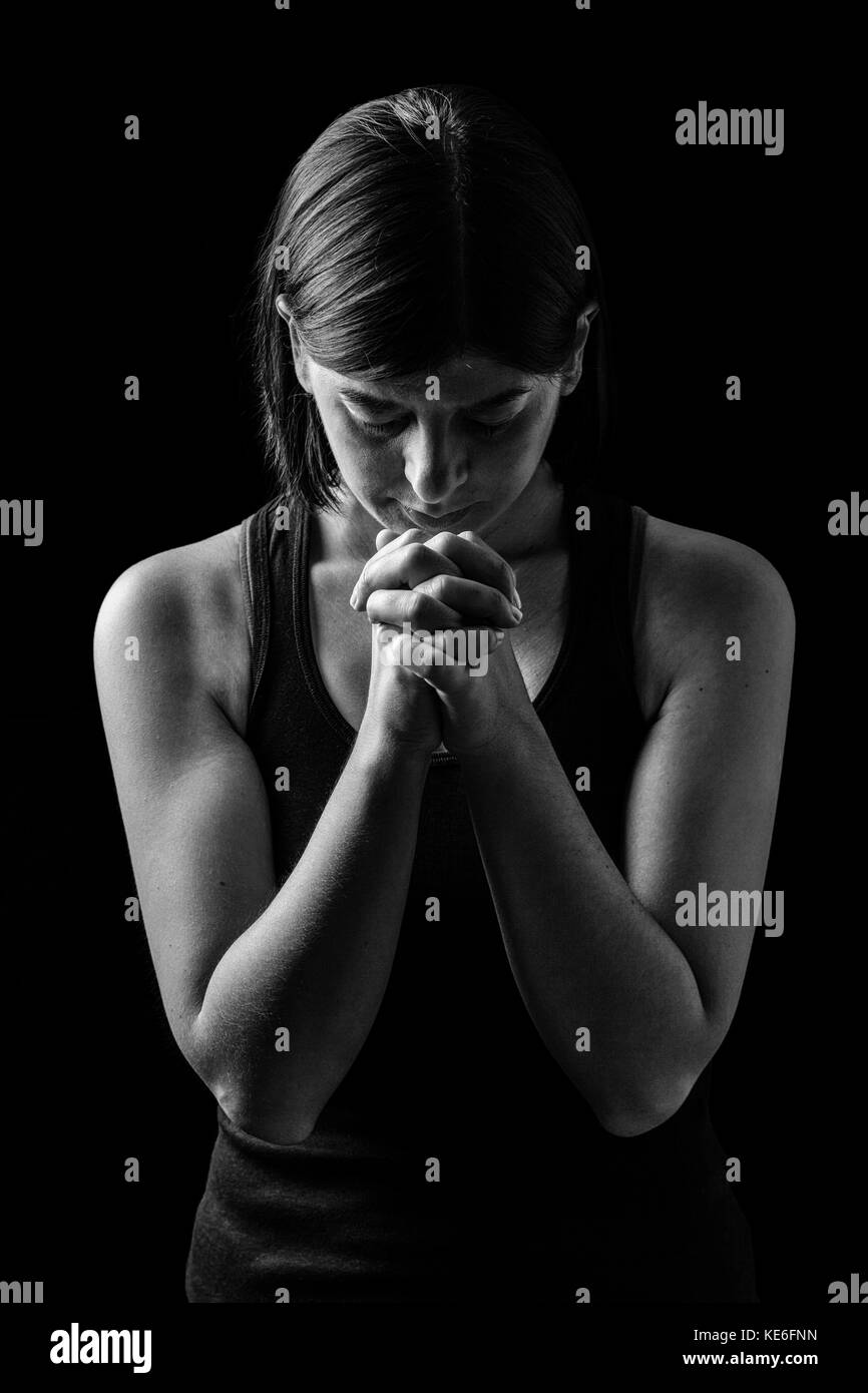 Faithful athletic woman praying, with hands folded in worship to god, head down and eyes closed in religious fervor, on low key black background. Stock Photo