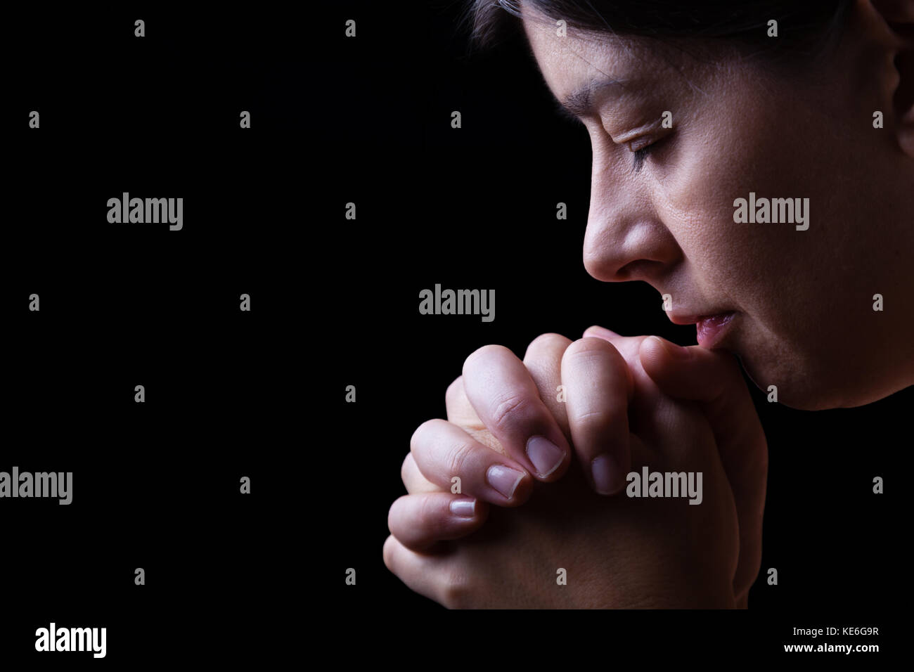 Faithful woman praying, hands folded in worship to god with head down and eyes closed in religious fervor, on a black background / prayer portrait Stock Photo