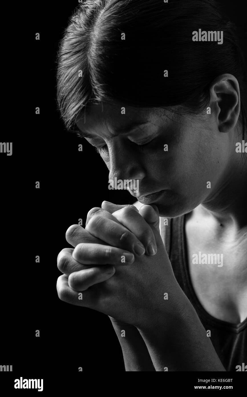 Low key portrait of a faithful woman praying, hands folded in worship to god, head down and eyes closed in religious fervor, on a black background. Stock Photo