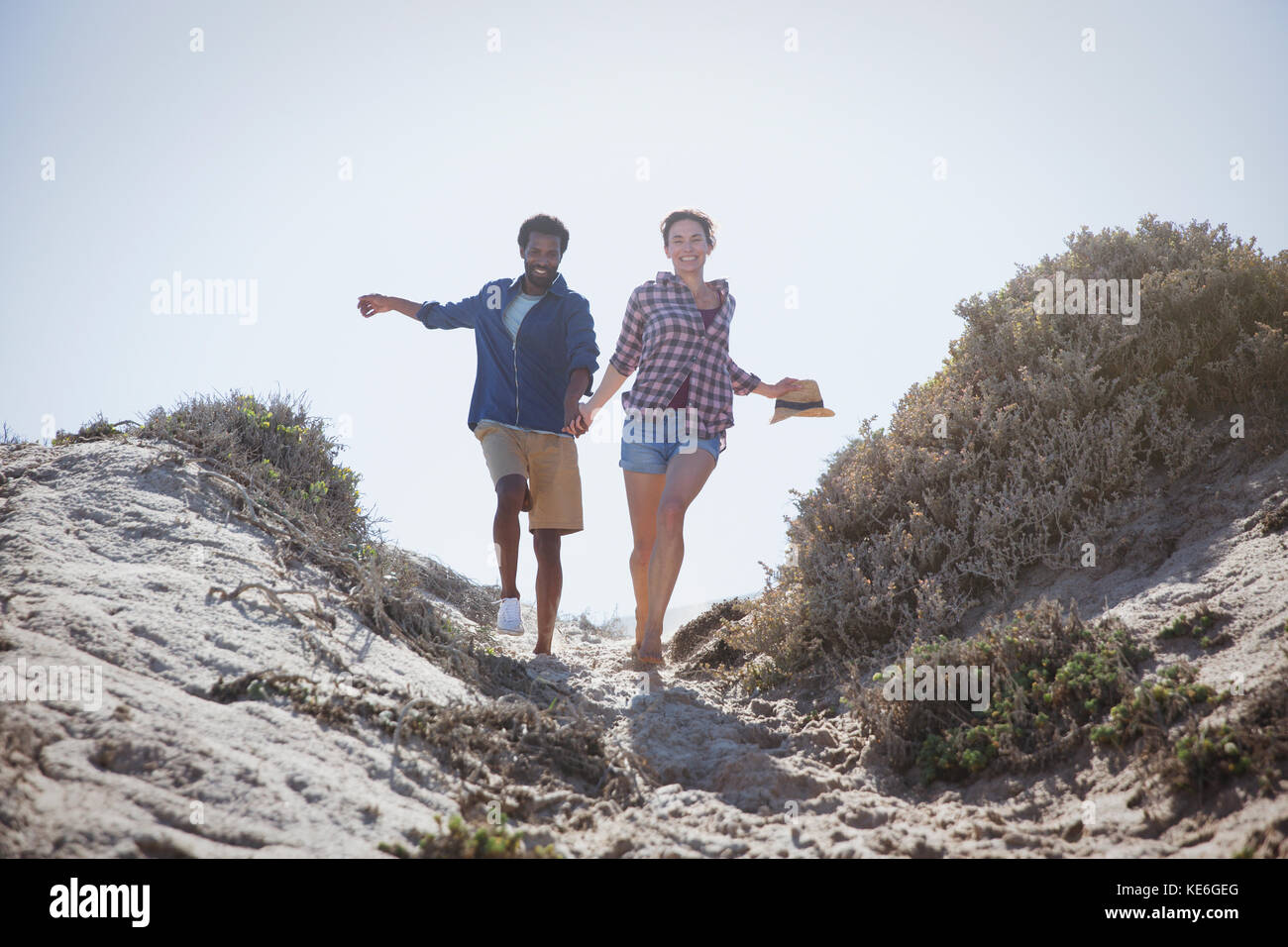 Playful, energetic multi-ethnic couple running and holding hands on sunny summer beach path Stock Photo