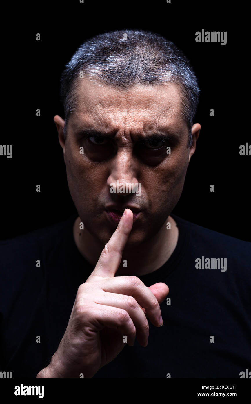 Furious mature man with aggressive look making silence sign in violent and threatening way. Black background / rage shh shhh shut up scary angry Stock Photo