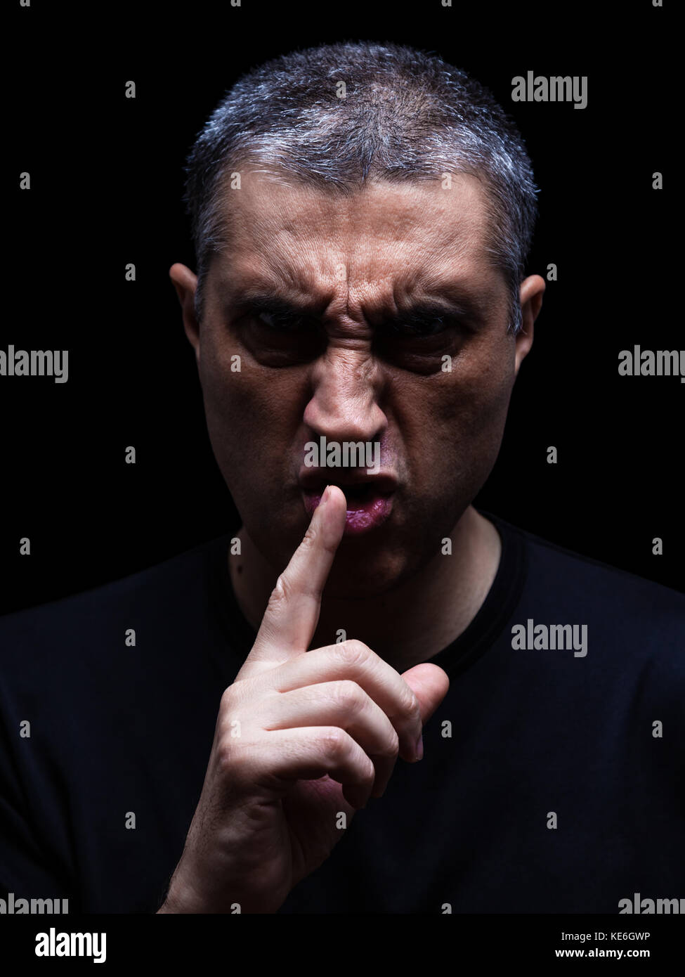 Furious mature man with aggressive look making silence sign in violent and threatening way. Black background / rage shh shhh shut up scary angry Stock Photo