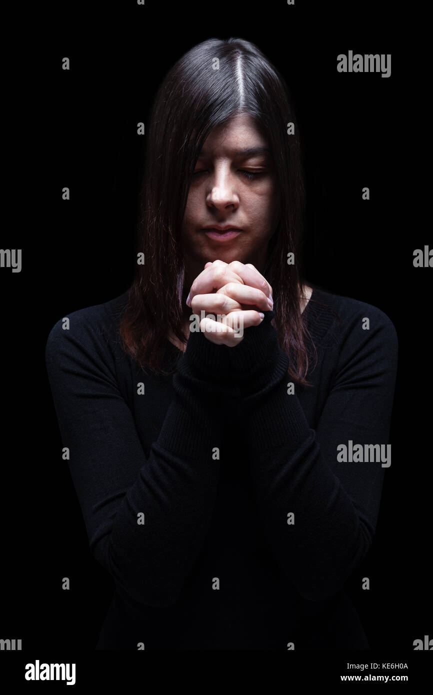 Mourning woman praying with hands folded in worship to god, looking down, eyes closed suffering. black background / faith prayer sad sadness mourning Stock Photo