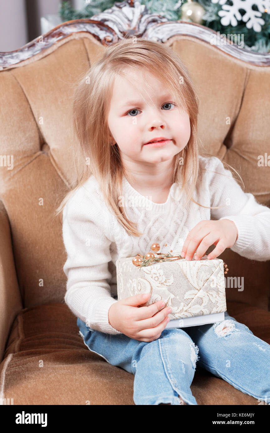 cute little girl sitting in a chair and opens a box with a present for ...