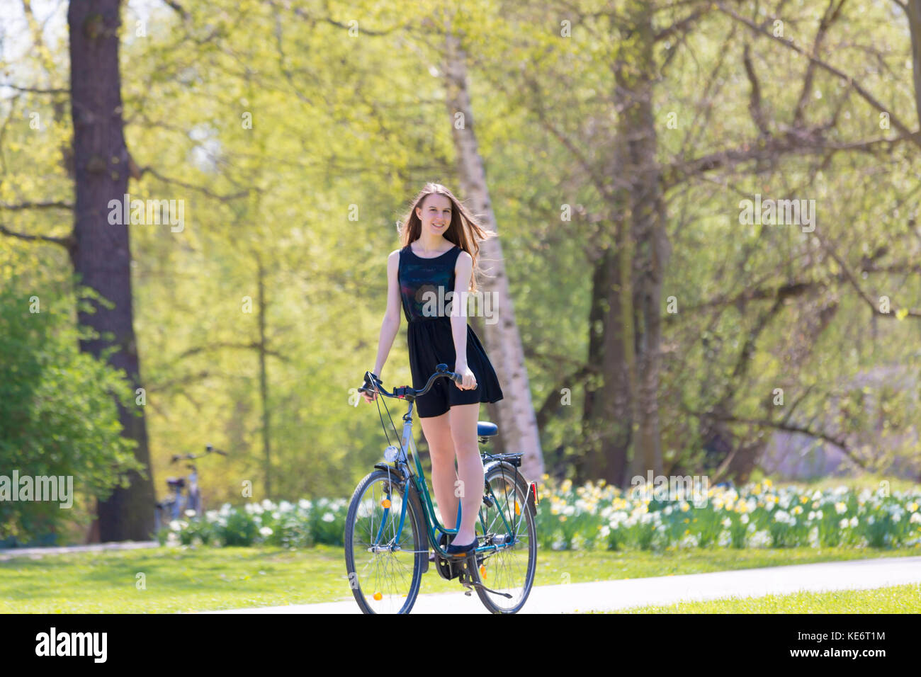 Portrait view of girl on bicycle wearing on black short dress. Young happy Woman riding along road on green spring  outdoor Park. Sporty young girl ri Stock Photo